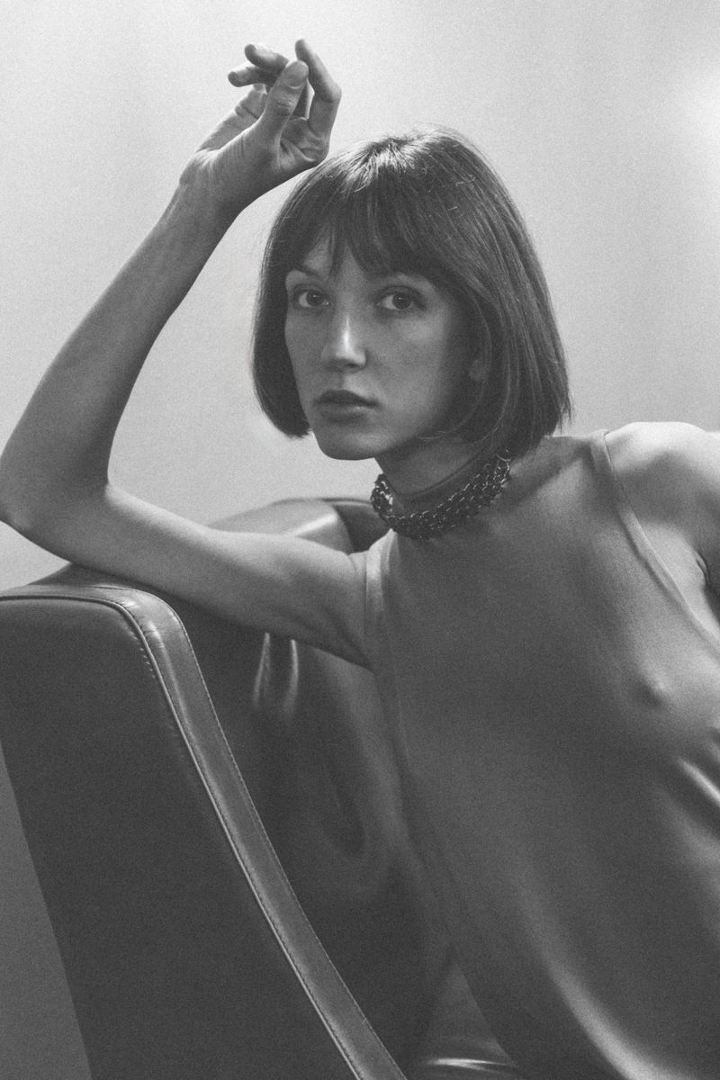 Monochrome portrait of a woman with short dark hair wearing a fitted top and choker, leaning on a leather chair.
