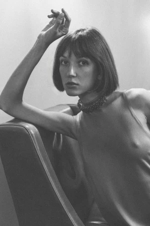 Monochrome portrait of a woman with short dark hair wearing a fitted top and choker, leaning on a leather chair.