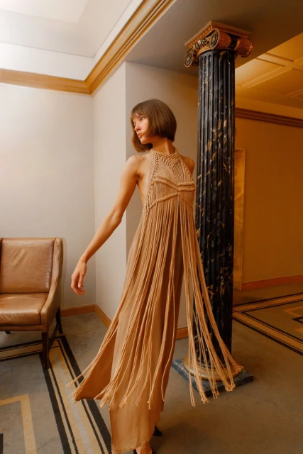 A slender adult woman poses indoors, wearing a light-brown macrame halter dress with long fringe, in a stylish interior with neutral tones.
