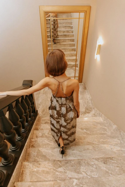 Woman in an evening dress walks down marble stairs with a wooden banister. The scene is reflected in a wall-mounted mirror.