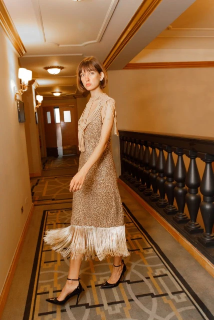A young woman walks through a hallway wearing a gold dress with a fringed hem and strappy heels, with golden light and neutral tones.