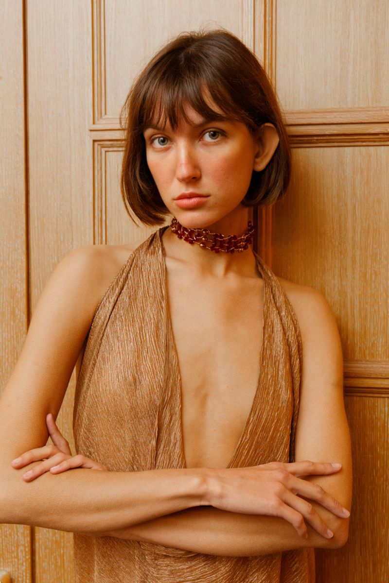 Indoor portrait of a woman in a gold dress with her arms crossed, featuring neutral makeup, a burgundy choker necklace, and a wood background.