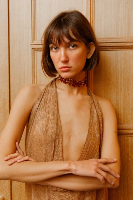 Indoor portrait of a woman in a gold dress with her arms crossed, featuring neutral makeup, a burgundy choker necklace, and a wood background.