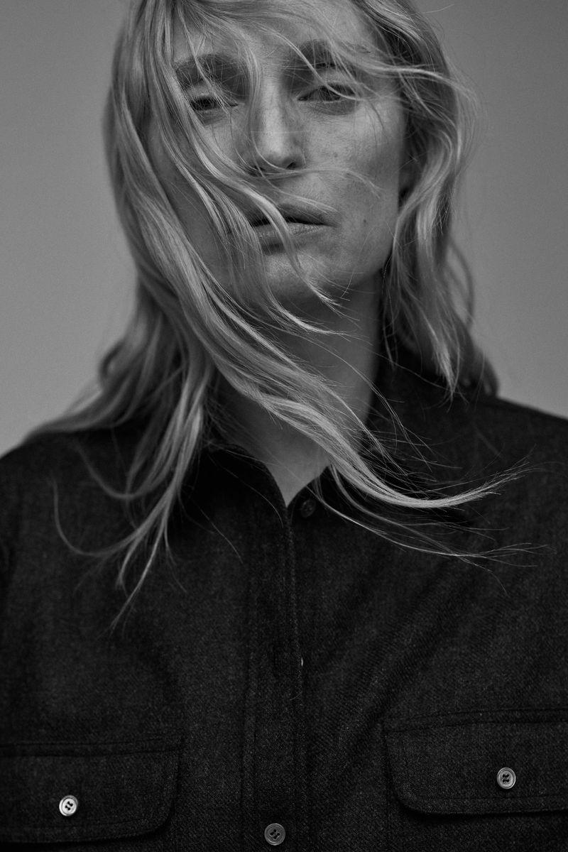 Portrait of a blonde woman with hair blowing across her face wearing a dark collared shirt against a grey background.