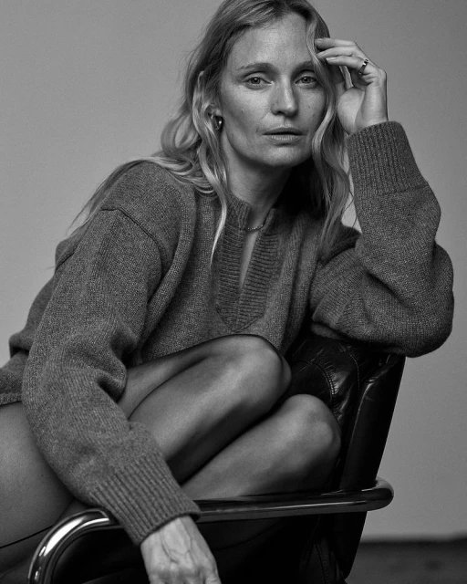 Black and white portrait of an adult woman with long blond hair sitting in a chair, wearing a sweater and black leggings.