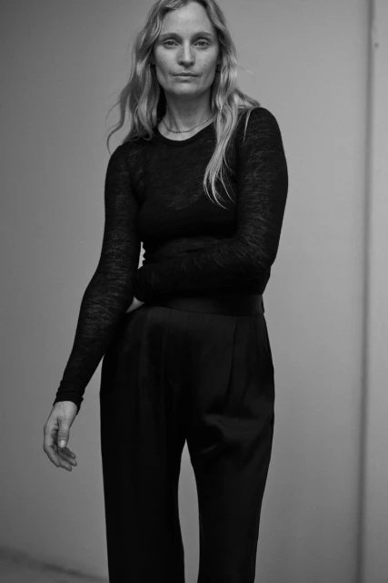 A woman in a long-sleeved black top and dark trousers poses in front of a neutral wall in a full shot, creating a minimalist fashion portrait.