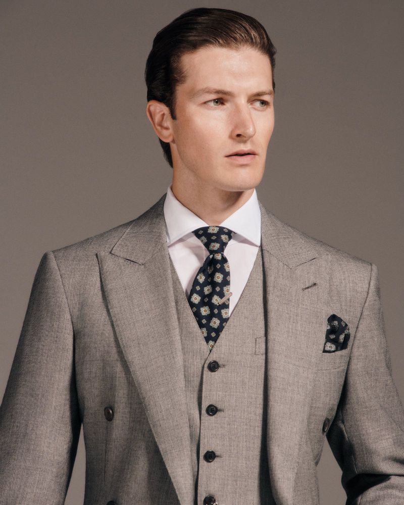 Man in a grey three-piece suit and patterned tie stands indoors against a grey background in a studio portrait, showcasing formal style.