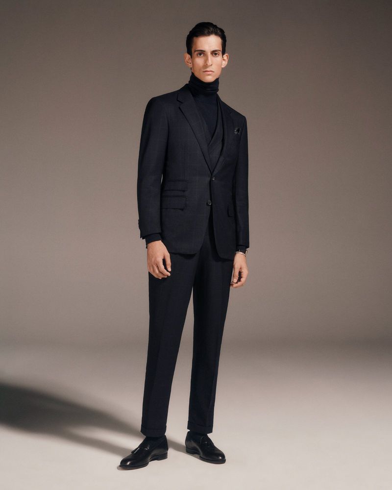 Studio shot of an adult male model wearing a navy blue three-piece suit with a turtleneck sweater and leather dress shoes.