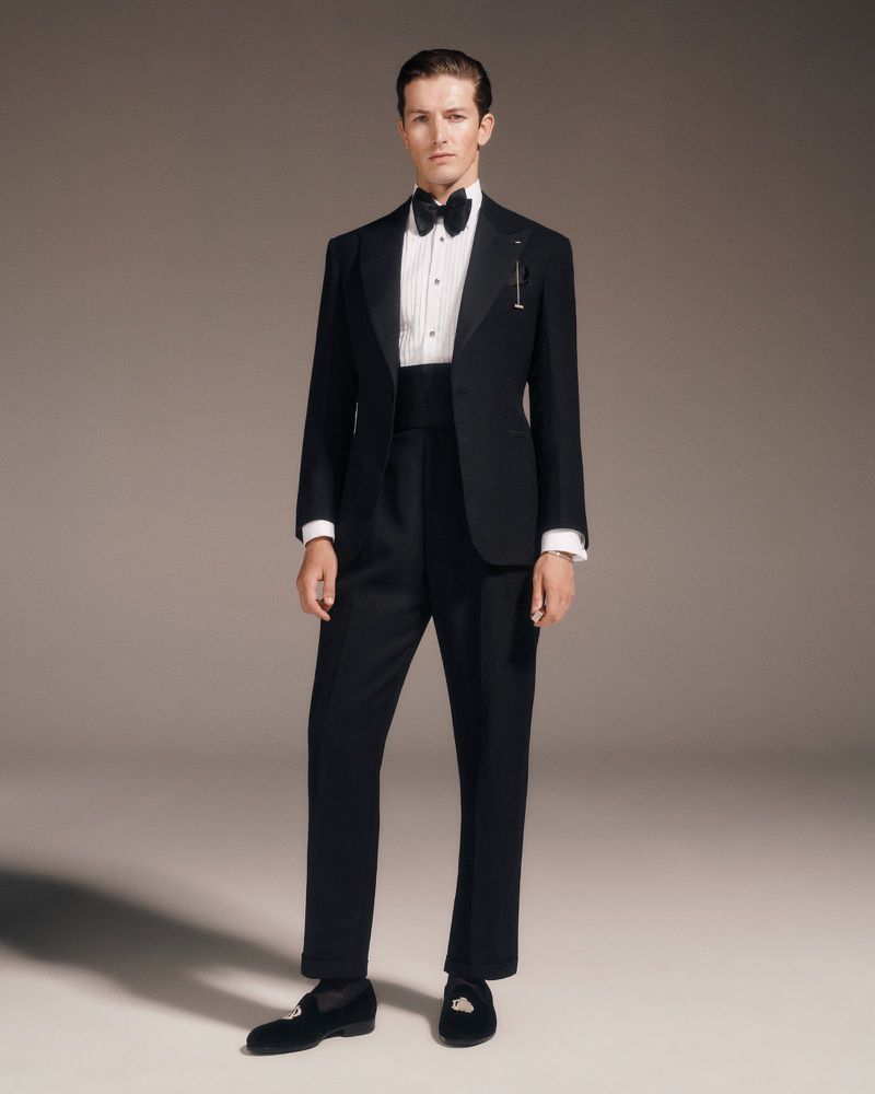 A man in a black tuxedo with a bow tie stands in a photo studio setting. He has dark shoes and a white shirt.