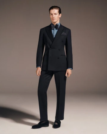 Full-length portrait of a man wearing a black tuxedo, bow tie, and dark dress shoes against a plain grey backdrop.
