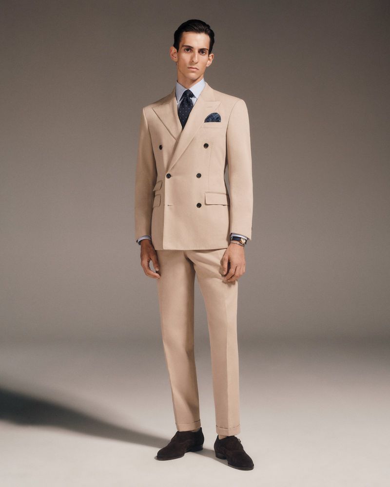 Man in a double-breasted tan suit standing indoors against a gradient background, styled with a blue shirt and tie, brown shoes.