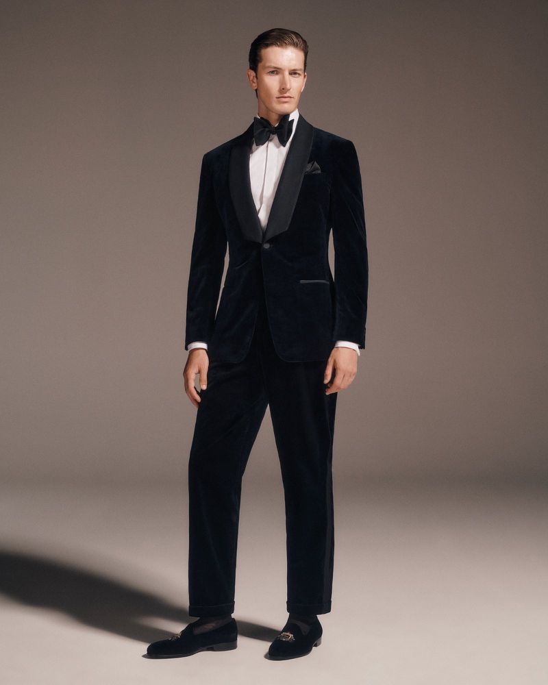 Full-length shot of an adult male in a formal dark-blue velvet tuxedo with a bow tie and loafers, standing against a neutral background.