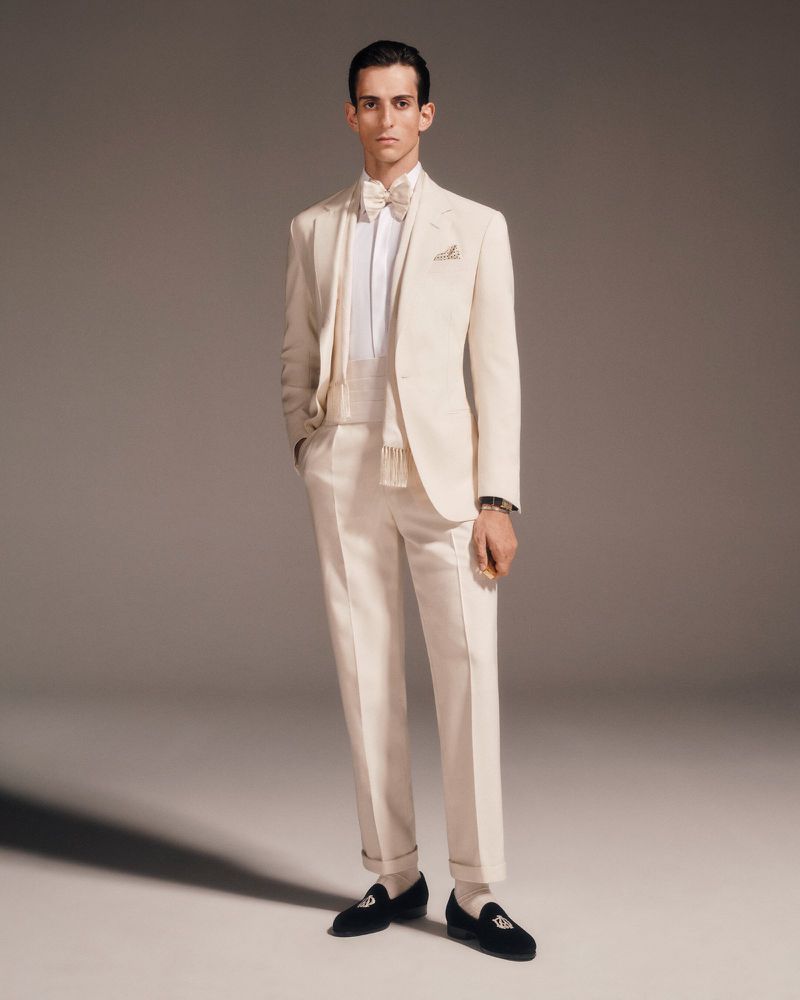 A man stands in a studio wearing a cream-colored formal suit with a bow tie and black loafers for a fashion shot with full lighting.