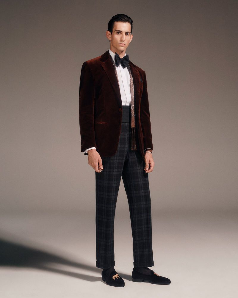 Man in a brown velvet suit and bow tie stands in a studio, posing for a full-length portrait with a neutral backdrop.