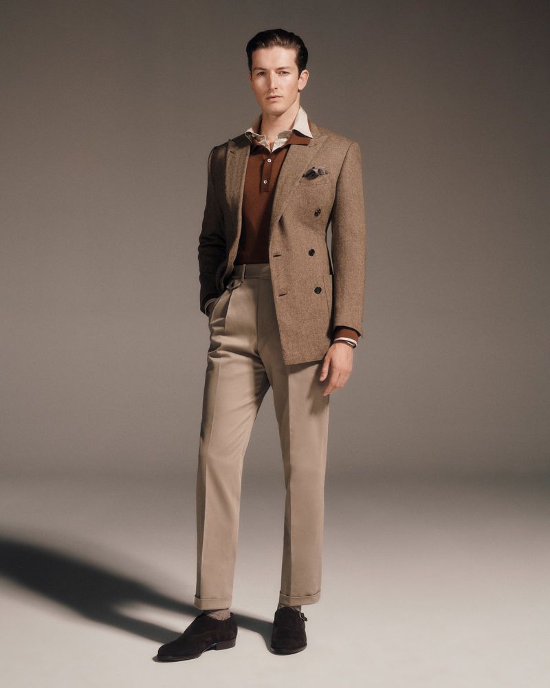 A full studio shot of a poised adult male, smartly dressed in a brown tweed blazer, collared shirt, and tailored trousers, set against a muted backdrop.