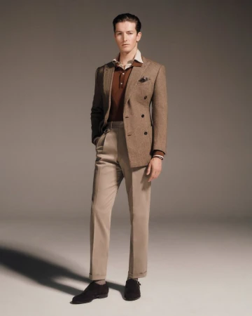A full studio shot of a poised adult male, smartly dressed in a brown tweed blazer, collared shirt, and tailored trousers, set against a muted backdrop.