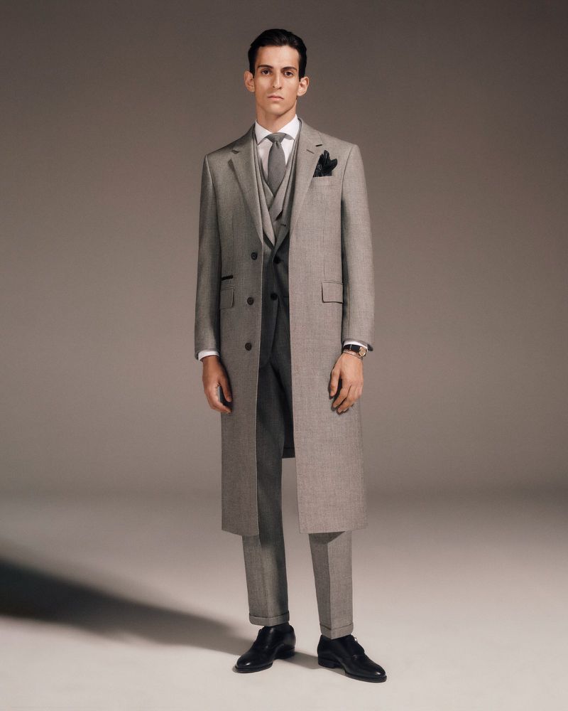 Full-length shot of a male model in a grey three-piece suit, grey overcoat, black shoes, and grey tie, set against a neutral backdrop.