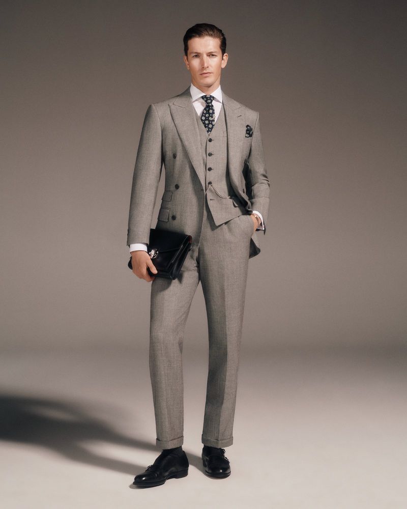 Man in a grey suit, vest, white shirt, and tie stands in a studio holding a black leather briefcase and wearing black dress shoes.