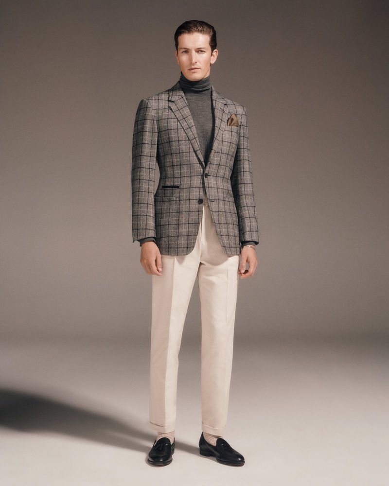 Man in a grey plaid blazer, turtleneck, and white pants, posing against a neutral background in a fashion editorial shot.