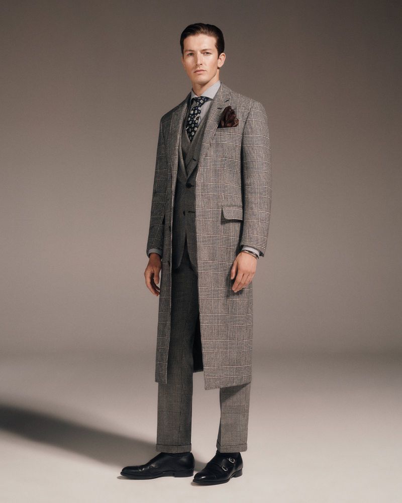 Full-length shot of a male model in a grey plaid overcoat, suit, tie, and black shoes against a neutral backdrop in a studio.