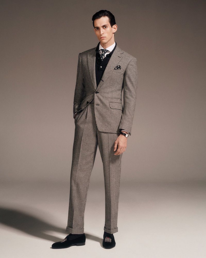 Portrait of a young adult male in a checkered suit, wearing a vest and tie, posing indoors in a fashion or editorial style photograph.