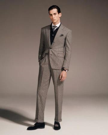 Portrait of a young adult male in a checkered suit, wearing a vest and tie, posing indoors in a fashion or editorial style photograph.