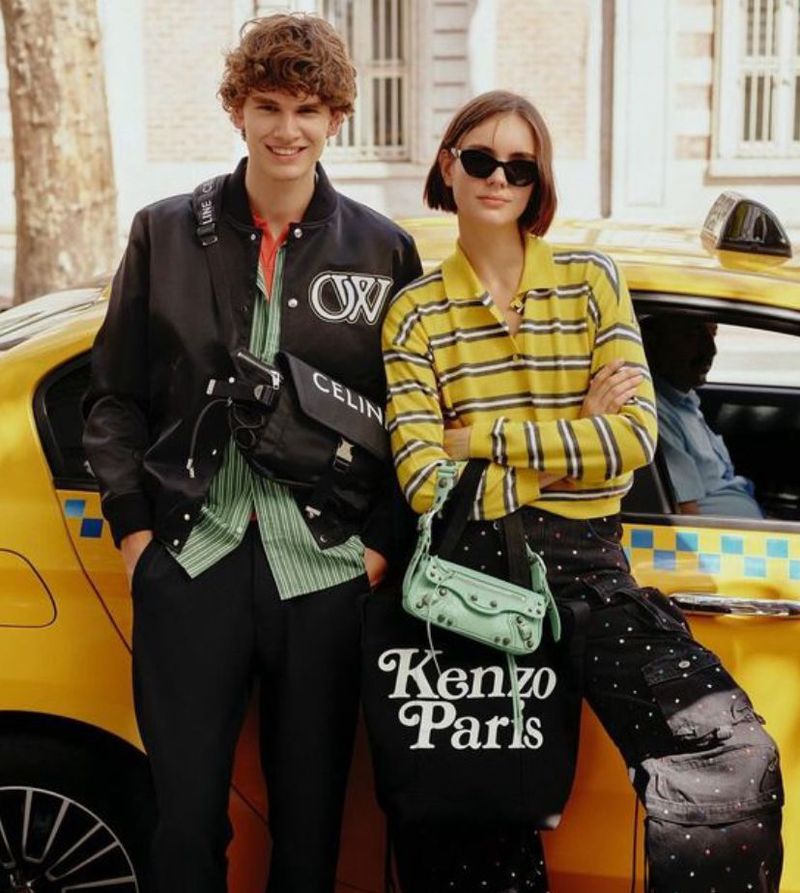 Two fashion models stand in front of a yellow taxi cab in an urban setting, showcasing clothing and accessories.