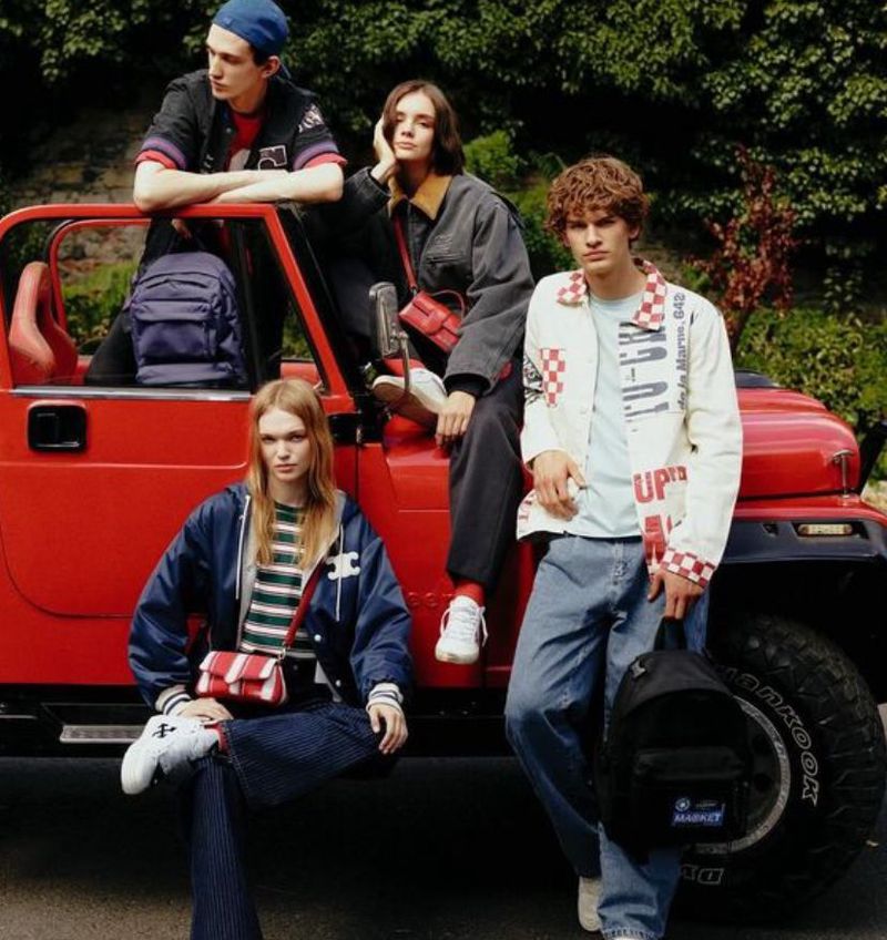 Group of four young adult fashion models pose alongside a red off-road vehicle wearing contemporary casual clothing and carrying backpacks.