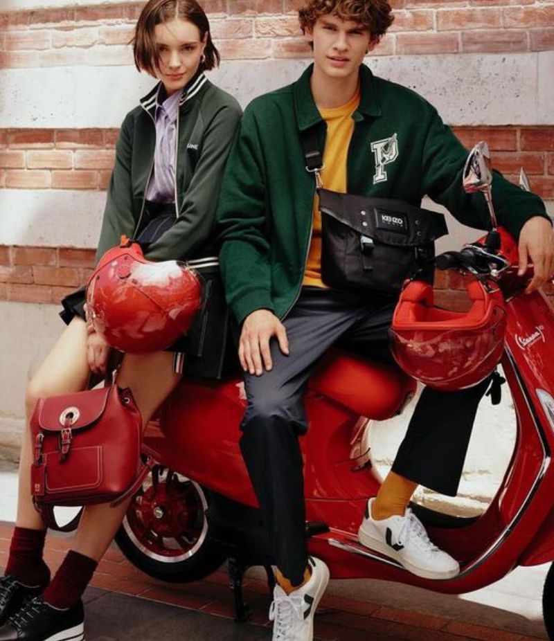 Two fashion models pose with a red Vespa scooter on a brick background, both wearing stylish green jackets and carrying red accessories.
