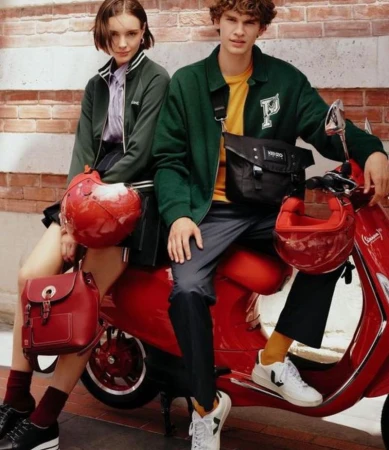 Two fashion models pose with a red Vespa scooter on a brick background, both wearing stylish green jackets and carrying red accessories.