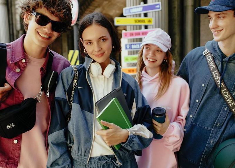 Four smiling students in casual clothing stand outdoors near colorful directional signs, holding books and a beverage, showcasing youthful style.