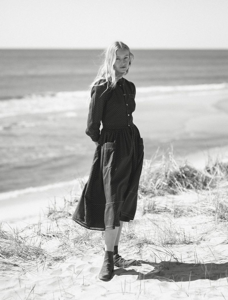 Black and white portrait of a woman in a dark dress standing on a sandy beach near the ocean on a sunny day with clear blue sky.
