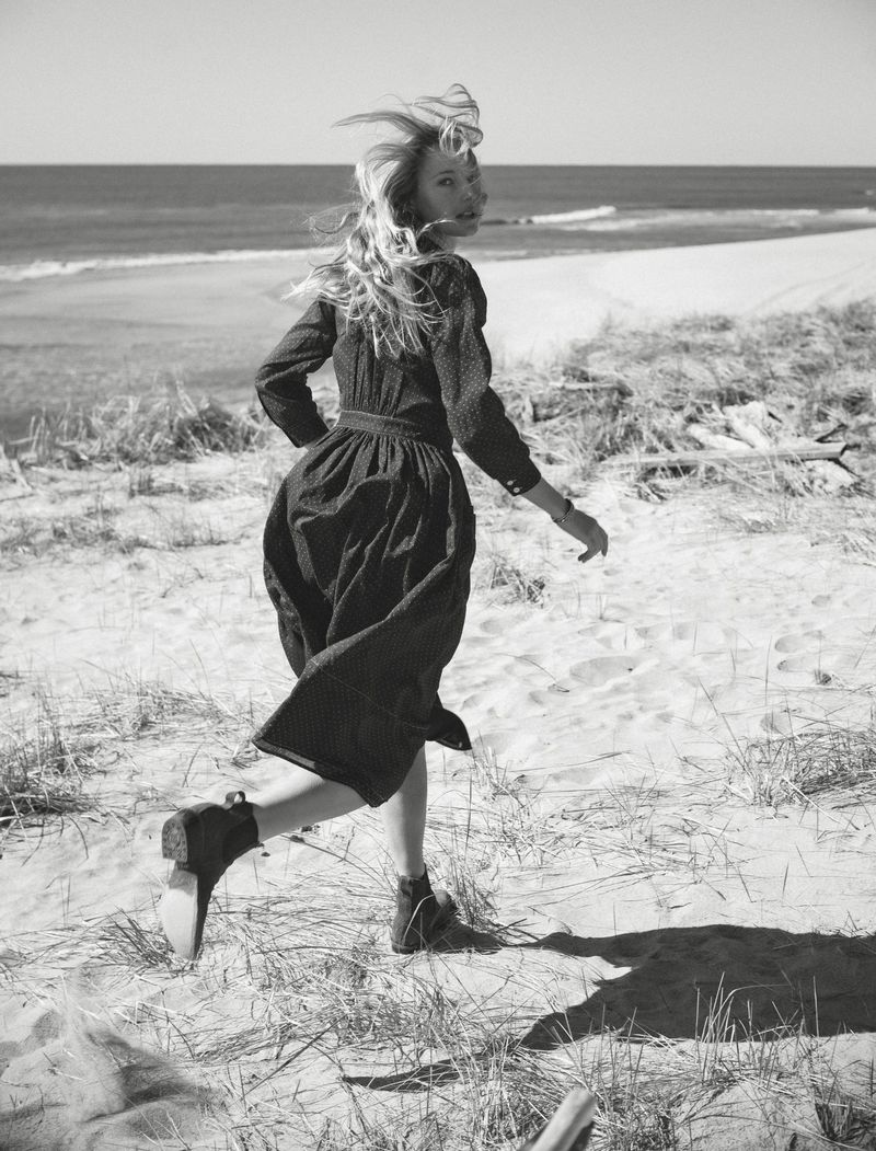 Black and white shot of a blonde female fashion model running along a sandy beach in a long-sleeved dress, looking back towards the camera.