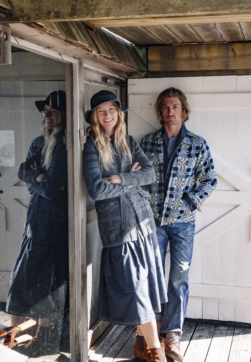 A smiling blonde woman in a grey cardigan and dark skirt and a man in a plaid jacket posing outdoors near a rustic wooden structure.