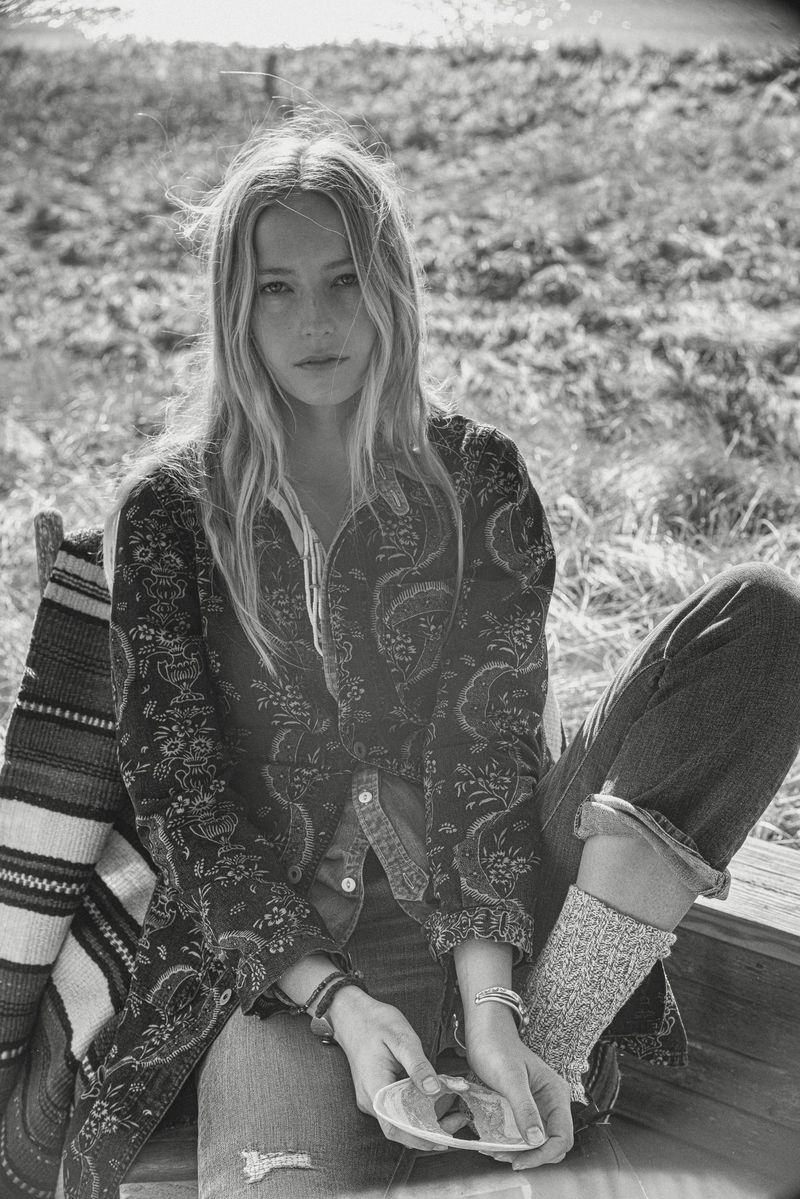 Black and white portrait of a woman sitting outside, wearing a patterned jacket, jeans, and knitted socks, holding a seashell.