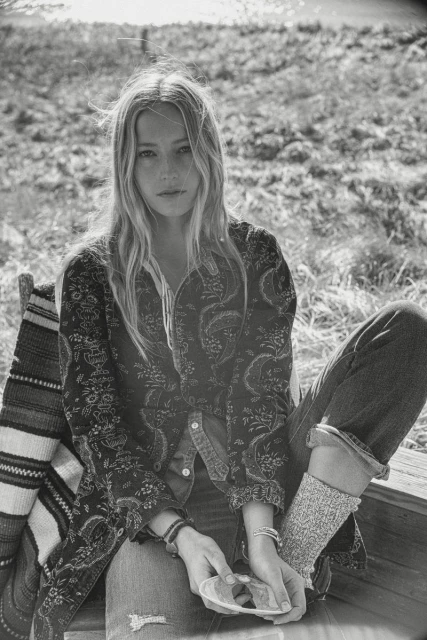 Black and white portrait of a woman sitting outside, wearing a patterned jacket, jeans, and knitted socks, holding a seashell.