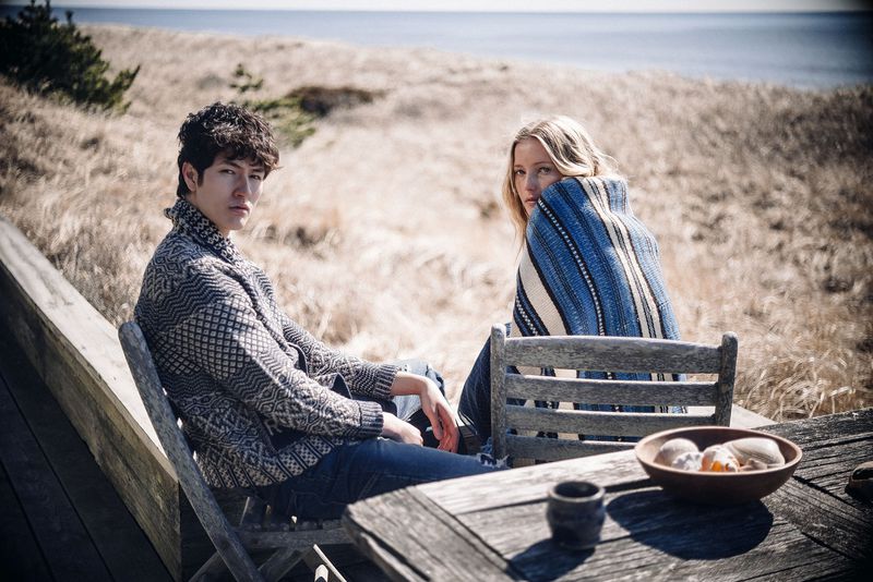 A young couple sitting outside on a wooden deck overlooking beach grass with the ocean in the background, wearing patterned sweaters and blankets.
