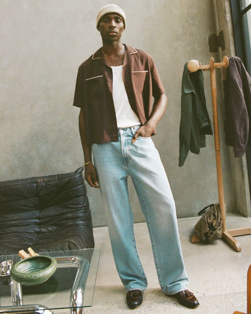 A tan-skinned male model in a brown short-sleeve shirt, white tank top, light blue jeans, and a tan beanie stands indoors.