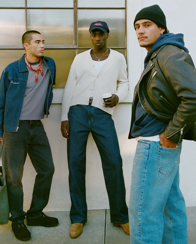 Three stylish men pose outdoors in casual denim and knitwear, standing against a textured light wall with natural light and shadows.