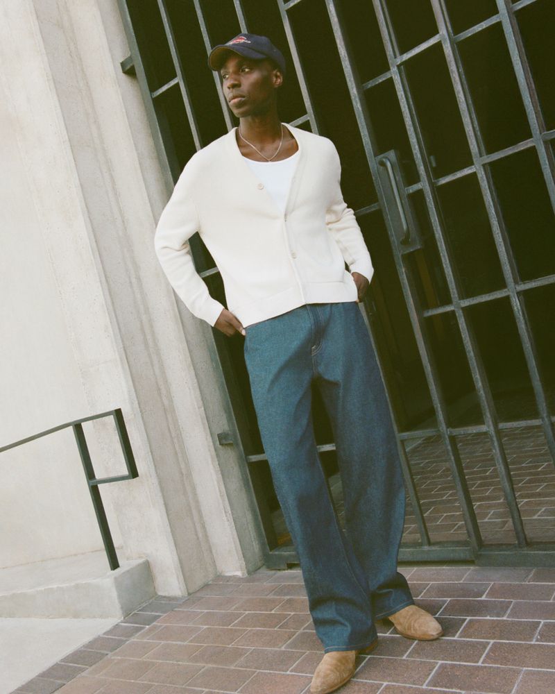 Male model stands against a gate wearing a cap, cream cardigan, t-shirt, dark wash jeans, and suede shoes in an urban environment.