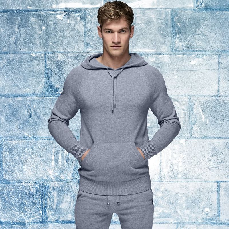 Adult male posing in a grey hooded sweatshirt and matching sweatpants against a faux icy blue brick-like background in a studio shot.