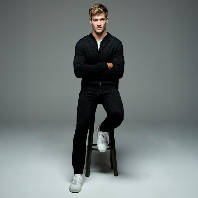 Studio portrait of a blond male model in a dark zip-up jacket and sweatpants, seated on a stool with arms crossed, wearing white sneakers.