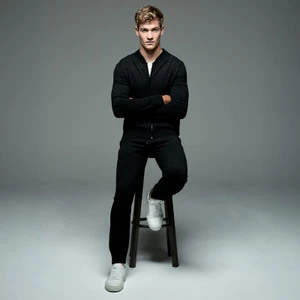Matty Carrington seated on a tall stool wearing a black knit zip jacket, black trousers, and white sneakers against a grey studio backdrop.
