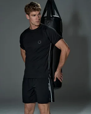 Male model wearing a black athletic t-shirt and shorts standing beside a dark punching bag against a grey seamless backdrop