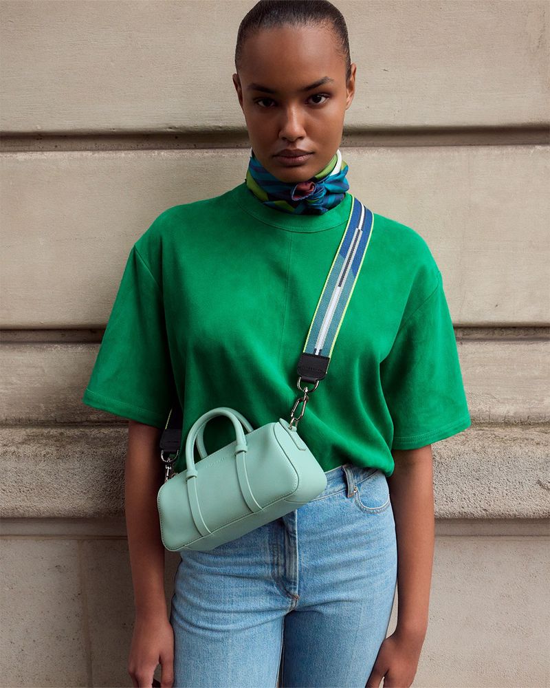 Woman in green t-shirt, light jeans, and a light-blue purse on a strap, poses against a light stone wall.