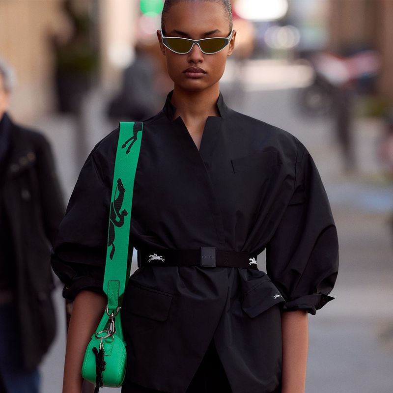 Stylish adult female with sunglasses in a black jacket and vibrant green bag outdoors, showcasing a fashion-forward, eye-catching ensemble.