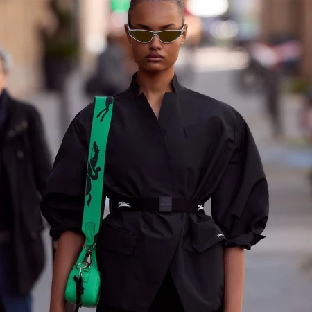 Stylish adult female with sunglasses in a black jacket and vibrant green bag outdoors, showcasing a fashion-forward, eye-catching ensemble.