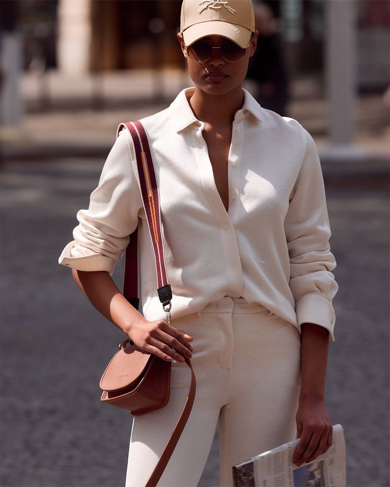 Woman in beige shirt, pants, and baseball cap stands outside with a brown cross-body purse and newspaper on a city street.