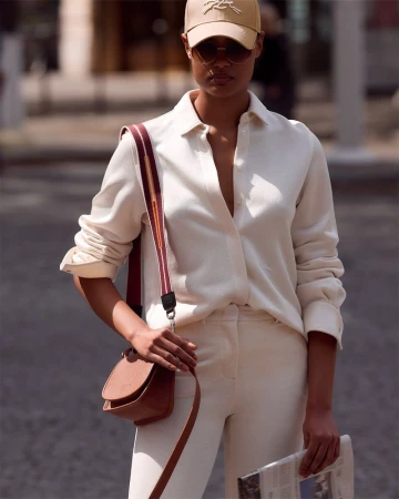 Woman in beige shirt, pants, and baseball cap stands outside with a brown cross-body purse and newspaper on a city street.