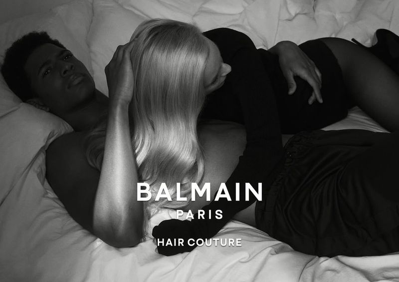 A couple lying in bed in a black and white fashion advertisement for Balmain Hair Couture. The pair are illuminated with studio lighting.